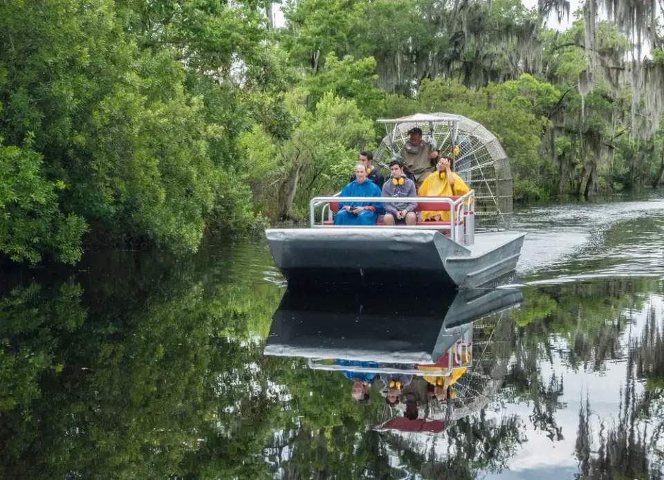 Airboat with poncho-clad passengers reflected on a calm swamp channel lined with mossy trees