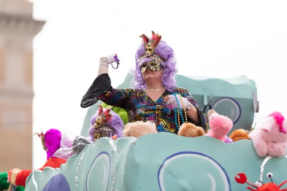 Costumed rider in purple wig and mask tossing beads and plush toys from a parade float