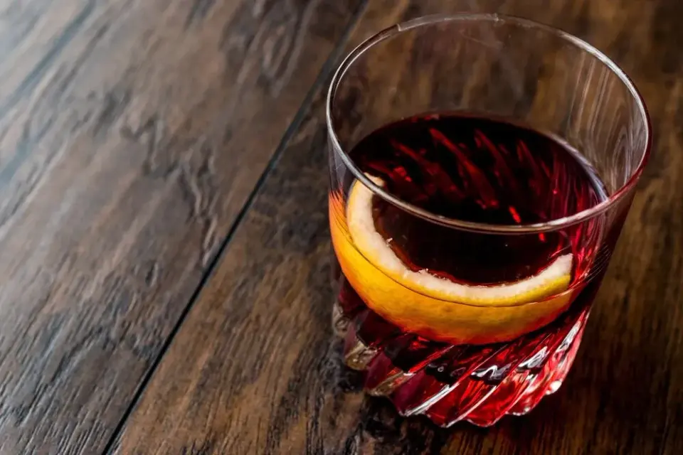Glass of red Sazerac-style cocktail garnished with a citrus twist on a dark wooden bar