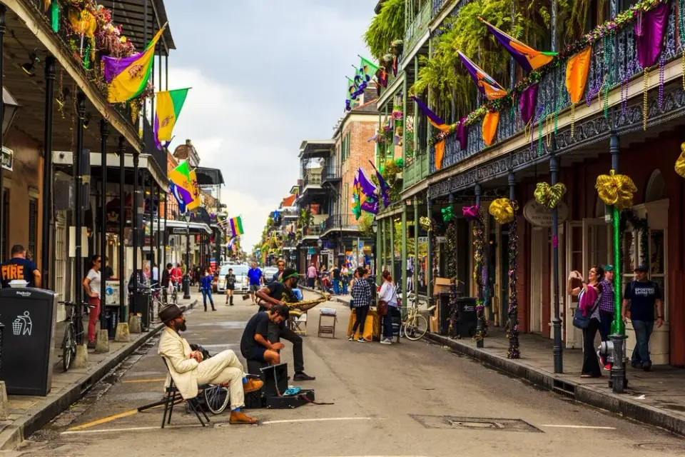 French Quarter street draped in purple, green, and gold Mardi Gras flags with a seated musician