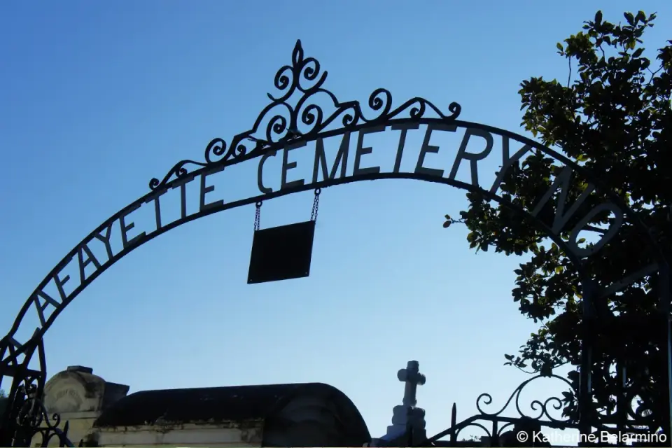 Wrought-iron Lafayette Cemetery archway silhouetted against blue sky above tombs and a cross