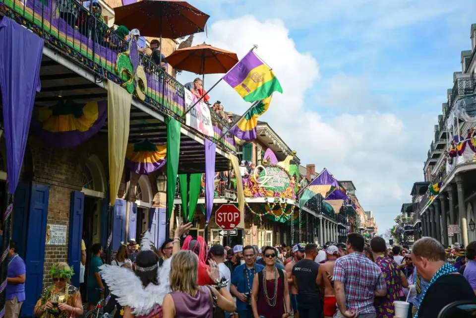 Crowded French Quarter street during Mardi Gras with purple, green, and gold flags and balcony revelers