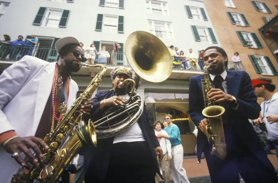 Three jazz musicians playing saxophones and sousaphone in a New Orleans street parade as onlookers watch