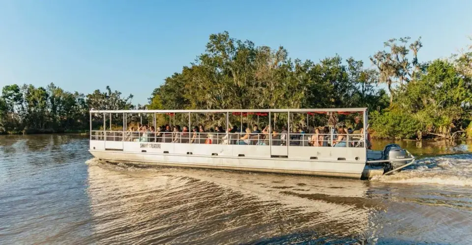 Covered swamp tour boat full of passengers cruising a sunlit bayou past wooded banks