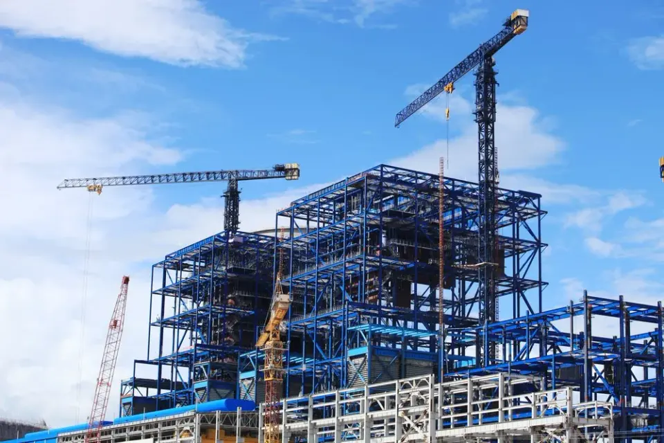 Steel-framed high-rise under construction with tower cranes against a bright blue sky