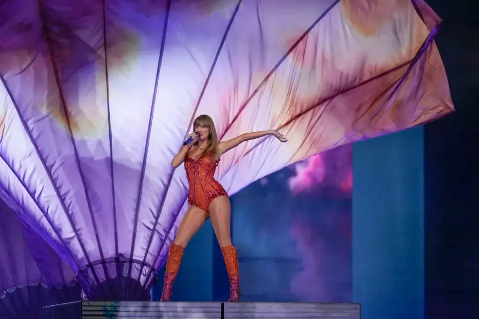 Taylor Swift singing on stage in a sparkling red bodysuit before a fan-shaped backdrop