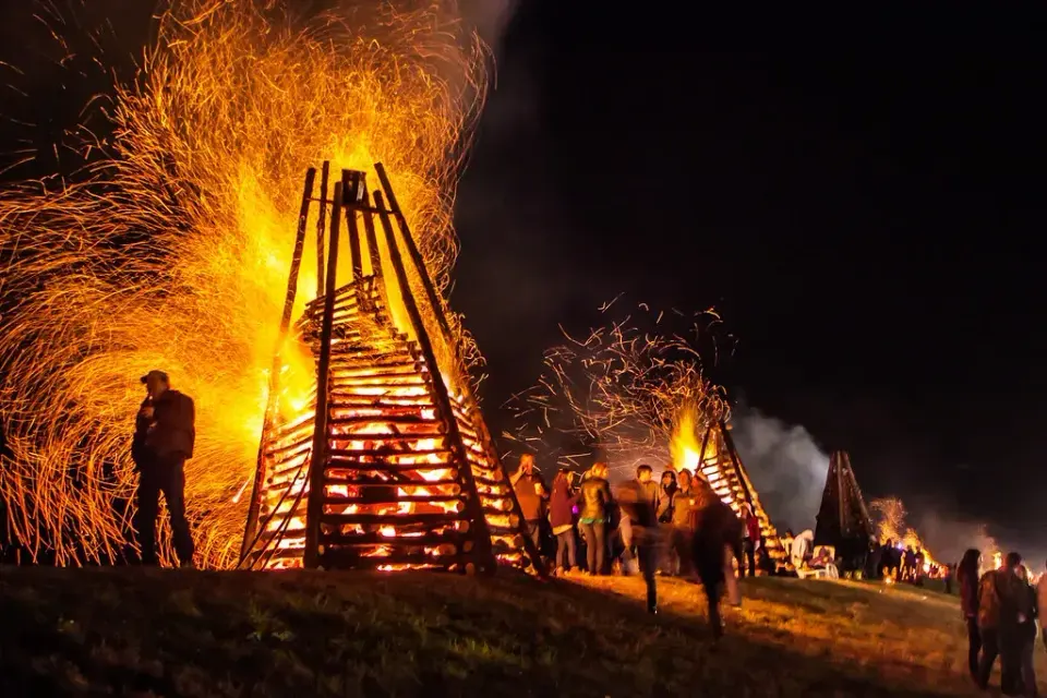 Towering wooden pyre bonfires blazing on the levee at night as crowds gather nearby