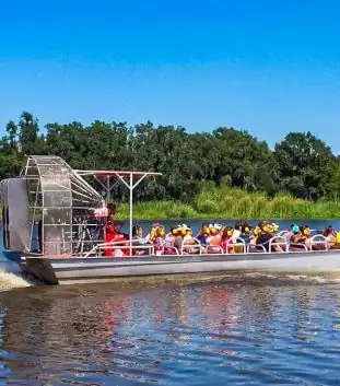 A large airboat filled with around 20 passengers skims across a broad Louisiana bayou under sunny skies during a New Orleans swamp tour.